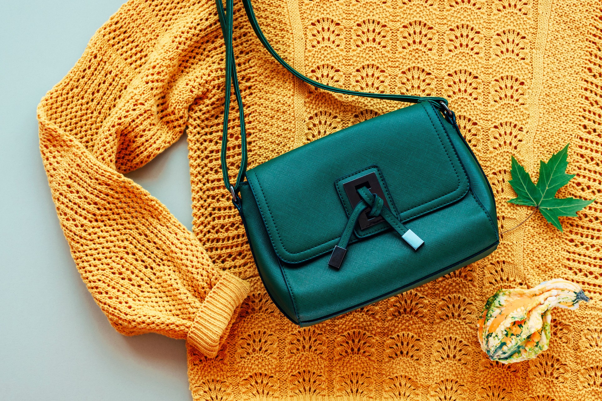 Flat lay of woman's yellow sweater and green bag with small pumpkin and autumn leaf on light green background. Autumn fashion concept. Top view, flat lay